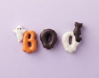 May include: The word BOO spelled out with three pretzels coated in icing. The B is orange, the O is dark brown, and the O is white. A small white ghost figure sits on the B, and a small dark brown cat figure sits on the final O.