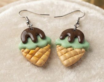 May include: A pair of dangle earrings featuring miniature ice cream cones with a textured tan waffle base, a scoop of light green mint-colored ice cream, and a glossy dark brown chocolate drizzle topping, suspended from silver-toned metal hooks.