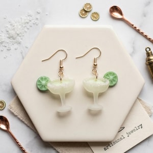 May include: A pair of margarita earrings with gold-colored hooks. Each earring features a miniature margarita glass with a lime wedge and a sugared rim. The earrings are displayed on a white hexagonal tile.