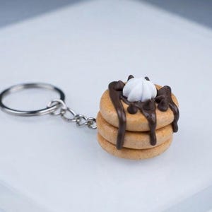 May include: A keychain featuring a miniature stack of three light brown pancakes topped with dark brown chocolate syrup, small chocolate chips, and a dollop of white whipped cream, attached to a circular silver metal split ring by a short chain.