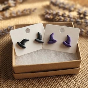 May include: Two pairs of stud earrings shaped like witch hats, one pair in solid black and one pair in solid purple, each mounted on a white rectangular display card and presented inside a small square cardboard box with white padding.