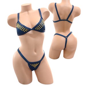 May include: A denim bikini set with gold-colored stud embellishments. The triangle top and thong-style bottoms are displayed on a mannequin. The bikini is a dark blue color.