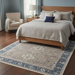 May include: A bedroom scene featuring a wooden bed frame, a beige patterned rug, and a matching beige quilt. The rug has a navy blue border and a central design in beige and blue. A beige armchair and a nightstand with a lamp are also visible.