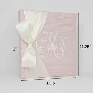 Pode incluir: Um livro de visitas de linho rosa com um la&ccedil;o de cetim branco e um monograma bordado branco. O livro mede 26,7 cm de largura por 28,6 cm de altura.