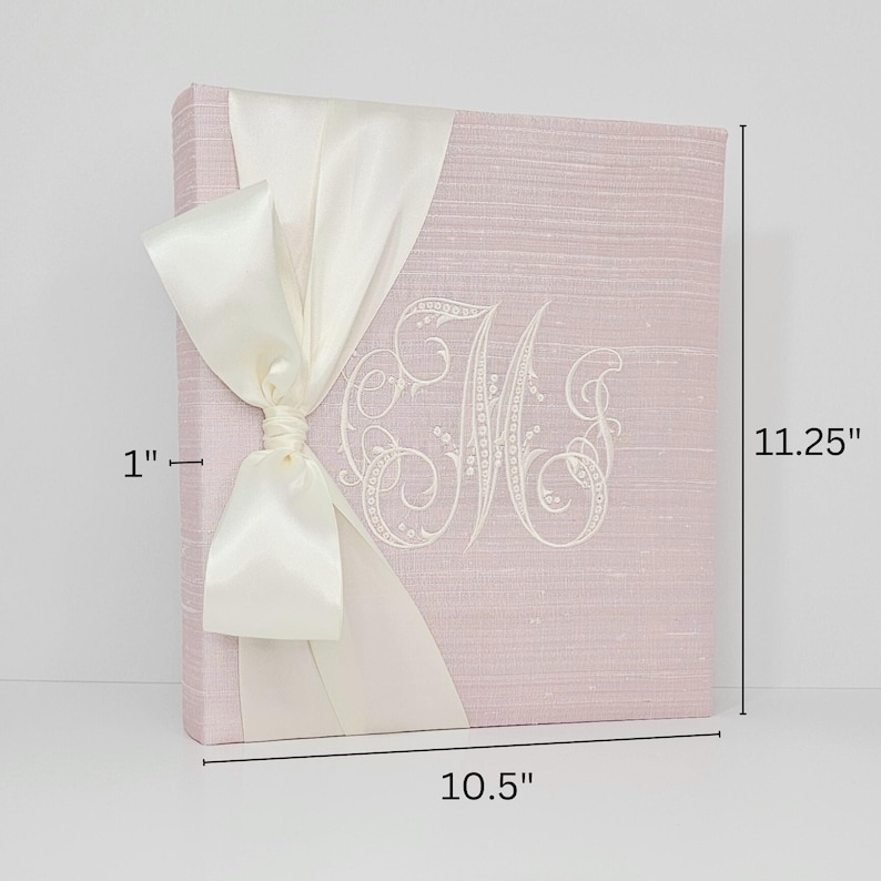 Pode incluir: Um livro de visitas de linho rosa com um la&ccedil;o de cetim branco e um monograma bordado branco. O livro mede 26,7 cm de largura por 28,6 cm de altura.