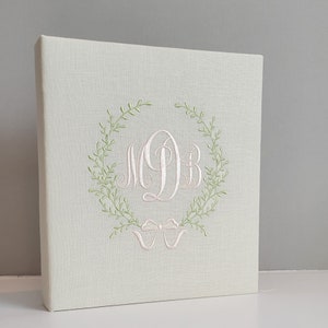 May include: A light beige fabric-covered photo album featuring an embroidered design on the front cover. The embroidery consists of a light green leafy wreath surrounding a large, white, cursive monogram of the letters MDB, with a white bow below.
