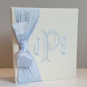 May include: A cream-colored photo album with a light blue ribbon and embroidered initials. The album features a textured cover and a decorative bow on the spine. The embroidered initials are in a light blue color.