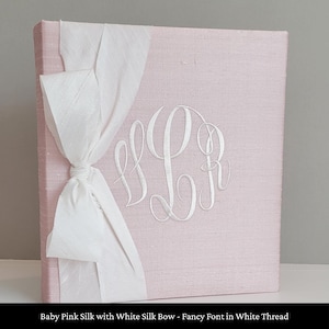 May include: A square photo album covered in baby pink silk fabric featuring a large white silk ribbon tied in a bow on the left side. The center of the cover displays a white embroidered monogram with the letters L and R in a fancy script font.
