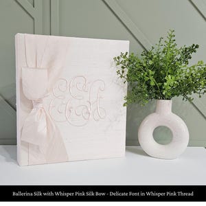 May include: A square, light pink fabric-covered album featuring a large, matching silk bow on the left side and an embroidered monogram with the letters E and H in the center, accompanied by a white, donut-shaped ceramic vase with green foliage.