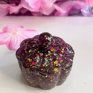 May include: A small, pumpkin-shaped trinket box. The box is dark with a glittery finish, featuring specks of pink, yellow, and green. The lid has a small, rounded handle. The background includes pink flowers.