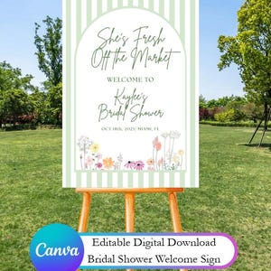 May include: A welcome sign for a bridal shower, displayed on a wooden easel outdoors. The sign features the text "She's Fresh Off the Market" and "Kaylee's Bridal Shower" with floral accents and the date. The sign also includes the text "Editable Digital Download".