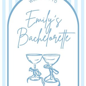 Pode incluir: Uma placa de boas-vindas listrada em azul claro e branco para a despedida de solteira de Emily. A placa apresenta um arco com o texto "Welcome to Emily's Bachelorette" e duas ta&ccedil;as de champanhe. A data e o local tamb&eacute;m est&atilde;o inclu&iacute;dos.
