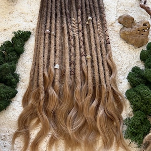 May include: A collection of brown and blonde dreadlocks with wavy ends. The dreadlocks are adorned with small beads and braided sections. The hair is displayed on a textured, off-white surface with green moss and wood accents.