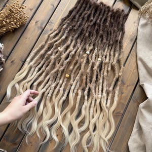 Może przedstawiać: Ombre dredloki w odcieniach brązu i blondu. Przedłużenia włosów ułożone są na drewnianej powierzchni, a dłoń delikatnie dotyka końców. Dredloki mają naturalny, teksturowany wygląd, niektóre zdobione są małymi koralikami.