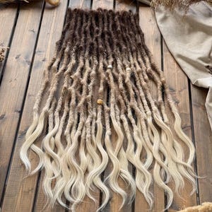 May include: A set of synthetic dreadlock extensions featuring a dark brown to light blonde ombre color gradient. The upper sections are textured dreadlocks, while the lower ends transition into loose, wavy, light blonde hair strands.