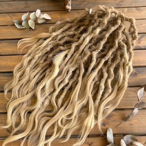 Puede incluir: Un conjunto de rastas rubias con textura ondulada, aseguradas con hilo blanco. Las extensiones de cabello están dispuestas sobre una superficie de madera, con ramas y hojas decorativas en el fondo.