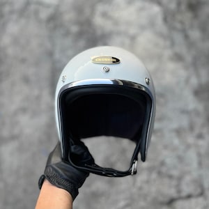 Pode incluir: Capacete de motocicleta cinza claro com acabamento cromado brilhante e interior preto. A palavra "CRUSHER" é exibida em uma placa dourada no capacete. O capacete é segurado por uma pessoa usando uma luva de couro preta.