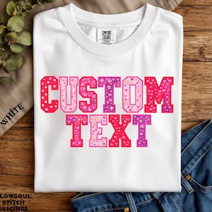 May include: A white short-sleeve t-shirt featuring the words CUSTOM TEXT in large block letters. Each letter is filled with a pink, red, and purple pattern of hearts, flowers, and small rainbows. The shirt has a crew neckline and a Comfort Colors label.