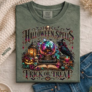 May include: A moss green t-shirt featuring a graphic print with the text "HALLOWEEN SPELLS" and "TRICK OR TREAT". The design includes a crystal ball, a raven wearing a witch hat, a glowing lantern, jack-o'-lanterns, skulls, and an open book.