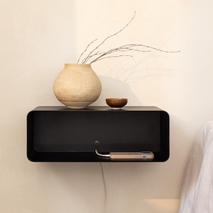 May include: A matte black rectangular floating wall shelf with rounded corners. On top sits a textured beige ceramic vase with thin branches and a small brown bowl. Inside the shelf, a smartphone is connected to a white charging cable.