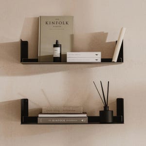 Può includere: Due mensole fluttuanti in metallo nero montate su una parete beige. La mensola superiore contiene un libro intitolato "THE KINFOLK GARDEN", una bottiglia nera e due libri bianchi. La mensola inferiore mostra libri e un diffusore nero.