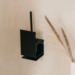 Può includere: Un portaspazzolino da toilette da parete in metallo nero. Il supporto ha una base rettangolare e un manico alto e sottile. Il design è minimalista e moderno. Il supporto è montato su una parete beige chiaro. Erba secca decorativa in un vaso bianco.
