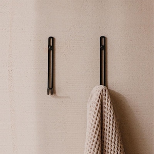 May include: Two vertical black metal wall hooks with a slender, elongated design and a small circular mounting point at the top. One hook is empty, while the other holds a beige waffle-textured fabric item draped over its lower end.