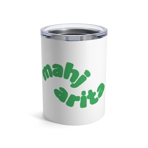 May include: White tumbler with a silver rim and lid. The tumbler features the green text "mahi arita" in a playful, curved font. The design is centered on the tumbler, making it a fun and eye-catching accessory.