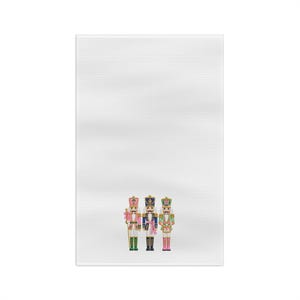 May include: A white rectangular kitchen towel featuring a printed graphic of three nutcracker figures standing in a row. The nutcrackers are dressed in colorful uniforms with pink, blue, green, and gold accents, each wearing a tall hat.