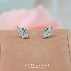 May include: A pair of silver stud earrings. Each earring has a wing design with small, sparkling stones and a pear-shaped, clear gemstone. The earrings are presented on a pink surface, with the text "BAREGEMGB Handcrafted Jewellery" at the bottom.