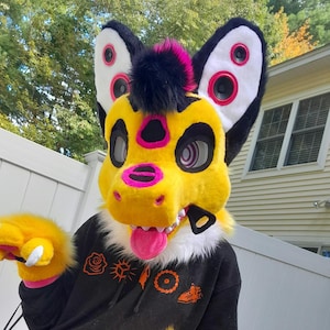May include: A yellow and black animal-inspired mascot head with a pink tongue, bright pink accents around the eyes and nose, and large ears featuring circular speaker-like details. The head has a black mohawk and is worn with a black hoodie.