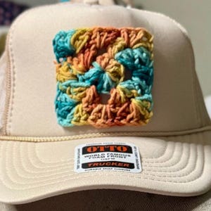 May include: A beige trucker hat featuring a square, multi-coloured crochet patch on the front panel. The patch displays a blend of teal, yellow, and orange yarn. A rectangular sticker on the brim reads OTTO WORLD FAMOUS FOAM FRONT OSFM TRUCKER.