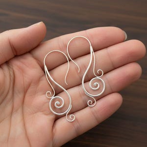 Könnte beinhalten: Ein Paar silberfarbene Drahtohrringe mit einem einzigartigen, wirbelnden Design. Die Ohrringe haben einen Haken für das Ohr und eine dekorative Spirale am unteren Ende. Die Ohrringe werden in einer Hand vor einem dunklen Holzgrund gehalten.