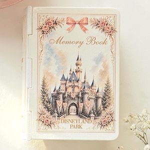 May include: A white rectangular memory book featuring a detailed illustration of a castle with the text "Memory Book" and "DISNEYLAND PARK" printed on the front cover, accented by a pink bow and floral borders in soft pastel tones.