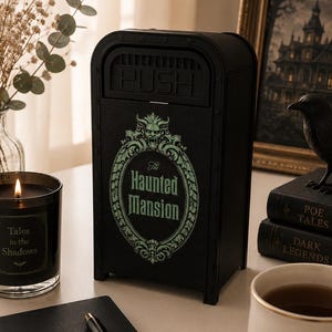 K&ouml;nnte beinhalten: Ein schwarzer Metall-M&uuml;lleimer mit gr&uuml;nem Haunted Mansion-Logo, eine schwarze Kerze mit der Aufschrift Tales in the Shadows, ein schwarzes Spiralnotizbuch mit goldenem Text FOCUS PLAN BLOOM, ein schwarzer Stift und eine wei&szlig;e Tasse mit goldener Schlossgrafik.