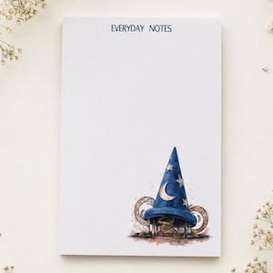以下が含まれることがあります： 上部に「EVERYDAY NOTES」と濃い青色で印刷された長方形の白いメモ帳。右下隅には、銀色の三日月と星で飾られた青い魔法使いの帽子の水彩画が描かれています。