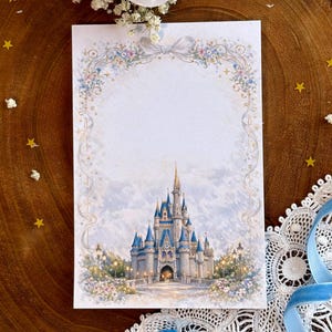 Könnte beinhalten: Aquarellillustration eines Märchenschlosses mit blauen Türmen und einer goldenen Spitze. Das Schloss ist von Blumen und Lichtern umgeben, vor einem weichen, bewölkten Himmel. Das Bild wird von einem floralen und Bandrahmen umrahmt.