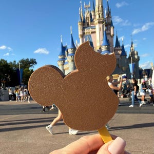 May include: A brown Mickey Mouse-shaped ice cream bar on a yellow stick, with a bite taken out of it. The background features a large castle with blue spires and a sunny sky, with people walking around.