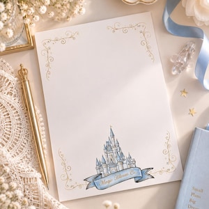 May include: A rectangular sheet of white stationery featuring gold filigree corner accents and a central illustration of a blue and white castle at the bottom. A blue banner draped across the castle base displays the text Magic Bloom Co.