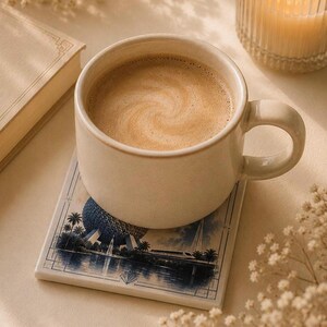 May include: A cream-colored ceramic mug filled with coffee sits on a square coaster featuring a blue and white illustration of a geodesic dome, palm trees, and a monorail. The mug has a smooth finish and a rounded handle, resting on a flat surface.