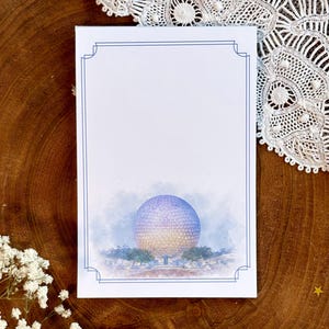 May include: A white card with a blue border features a watercolor illustration of a large, spherical structure, possibly a landmark. The image is set against a soft, blue and yellow background, with trees and a light blue sky. The card is on a wooden surface.