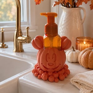 May include: An orange soap dispenser holder shaped like a pumpkin with mouse ears, featuring a carved smiling face and a base decorated with smaller pumpkins. The holder contains a clear plastic bottle with an orange pump mechanism on top.