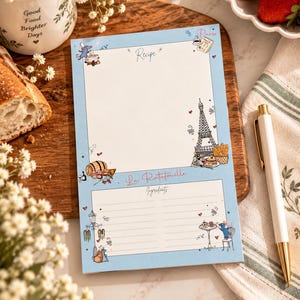 May include: A light blue rectangular recipe notepad featuring illustrations of the Eiffel Tower, a rat, and food items. The page includes the text Recipe, La Ratatouille, and Ingredients, with lined sections for writing down cooking instructions.