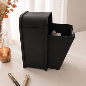 May include: A matte black desktop organizer featuring a tall, enclosed compartment with a curved top and an adjacent open-front bin. The structure is accented with vertical side panels secured by visible rivets along the edges, creating a sturdy look.