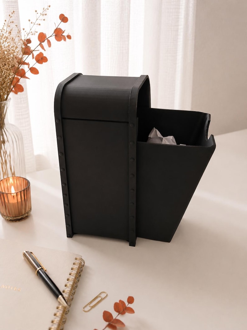 May include: A matte black desktop organizer featuring a tall, enclosed compartment with a curved top and an adjacent open-front bin. The structure is accented with vertical side panels secured by visible rivets along the edges, creating a sturdy look.
