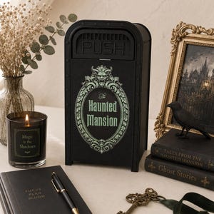 K&ouml;nnte beinhalten: Ein schwarzer, m&uuml;lltonnenf&ouml;rmiger Beh&auml;lter mit dem Schriftzug The Haunted Mansion in einem dekorativen ovalen Rahmen, einem gr&uuml;nen Wasserspeier-Akzent oben und einem PUSH-Etikett auf dem Klappdeckel, gestaltet mit dunkler &Auml;sthetik und Nieten.