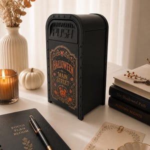 May include: A black metal mailbox with a rounded top and a hinged door labeled PUSH. The front features a decorative graphic with the text HALLOWEEN ON MAIN STREET, surrounded by orange pumpkin illustrations and intricate gold-toned scrollwork.
