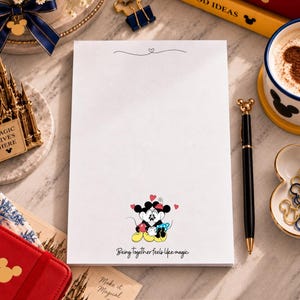 May include: A white rectangular notepad featuring a small illustration of Mickey and Minnie Mouse kissing at the bottom, surrounded by red hearts. The text "Being together feels like magic" is printed below the characters in a black script font.