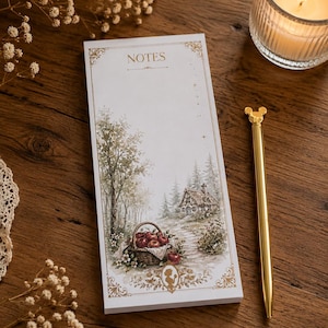 May include: A notepad with a landscape illustration featuring a cottage, trees, and a basket of apples. The notepad has the word "NOTES" at the top and is accompanied by a gold pen and a lit candle in a glass holder. The background is a wooden surface.