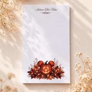 May include: A rectangular notepad with a white background featuring the text Autumn Glow Notes at the top. The bottom section displays a central jack-o'-lantern with two smaller pumpkin ears, surrounded by a cluster of dark orange autumn leaves.